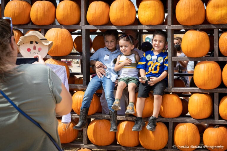 Three young boys pose for a photo while sitting on a wooden pumpkin display at a pumpkin patch, as a woman takes their picture and a scarecrow stands nearby.