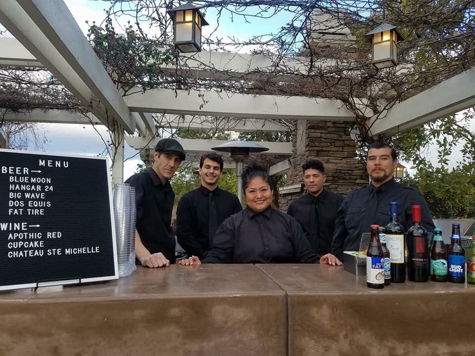 Five people stand behind an outdoor bar. A drink menu and various beverage bottles are displayed on the counter.