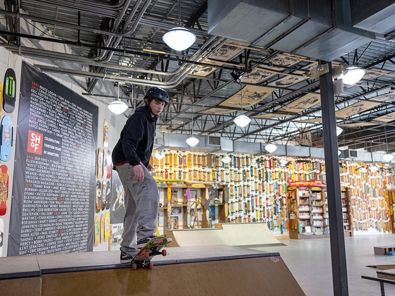 A person in a helmet skateboards indoors on a ramp, surrounded by walls decorated with skateboards and a large text mural.